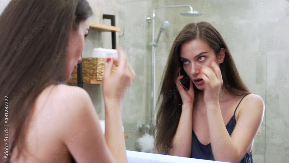 Pretty brunette woman in nigth clothes is looking at herself in the bathroom mirror and talking on the phone with a friend. Morning routine Pretty brunette woman in nigth clothes is looking at herself in the bathroom mirror and talking on the phone with a friend. Morning routine