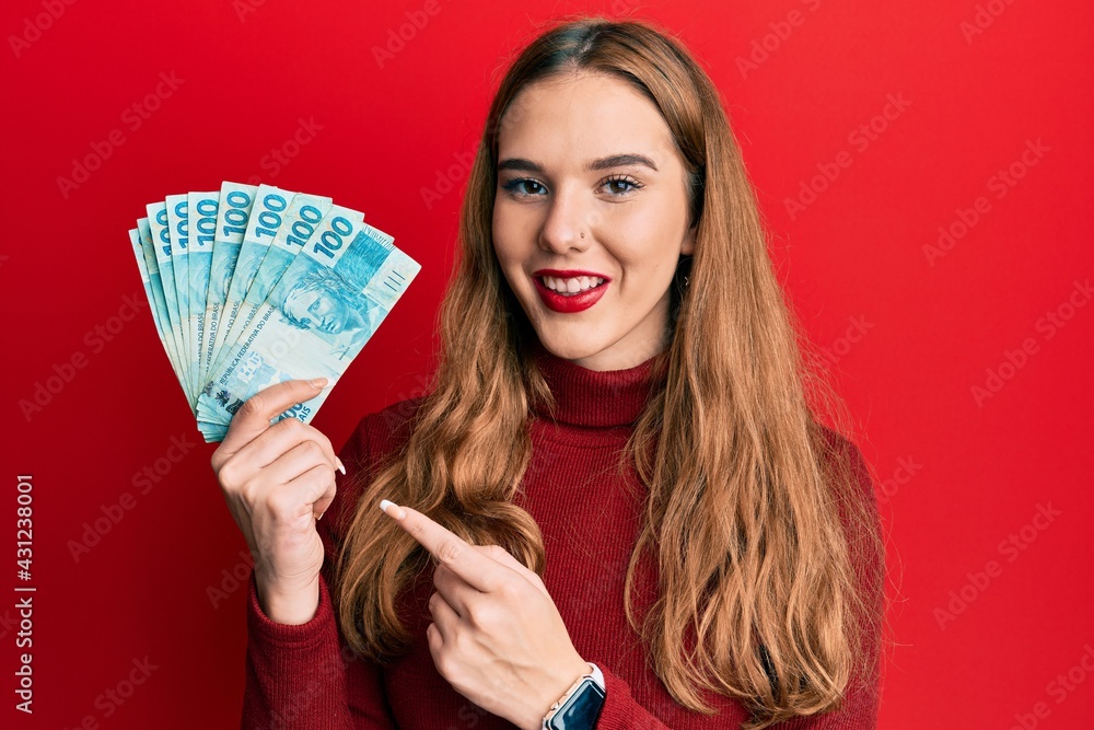 Young blonde woman holding 100 brazilian real banknotes smiling happy pointing with hand and finger