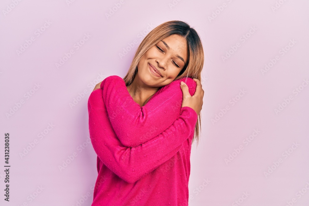 Beautiful hispanic woman wearing casual pink sweater hugging oneself happy and positive, smiling confident. self love and self care Beautiful hispanic woman wearing casual pink sweater hugging oneself happy and positive, smiling confident. self love and self care