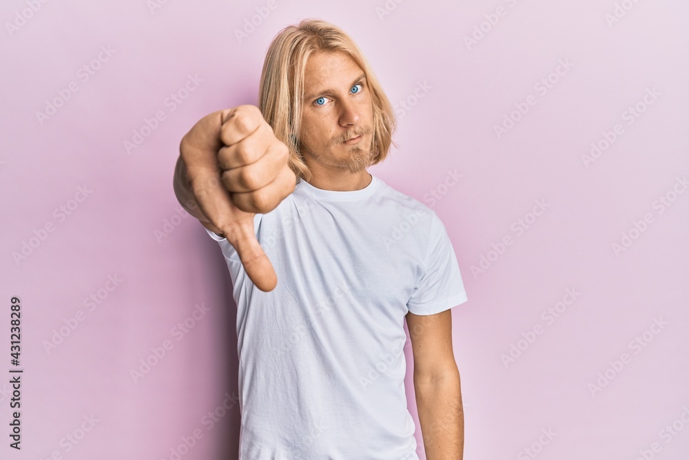 Caucasian young man with long hair wearing casual white t shirt looking unhappy and angry showing rejection and negative with thumbs down gesture. bad expression. Caucasian young man with long hair wearing casual white t shirt looking unhappy and angry showing rejection and negative with thumbs down gesture. bad expression.