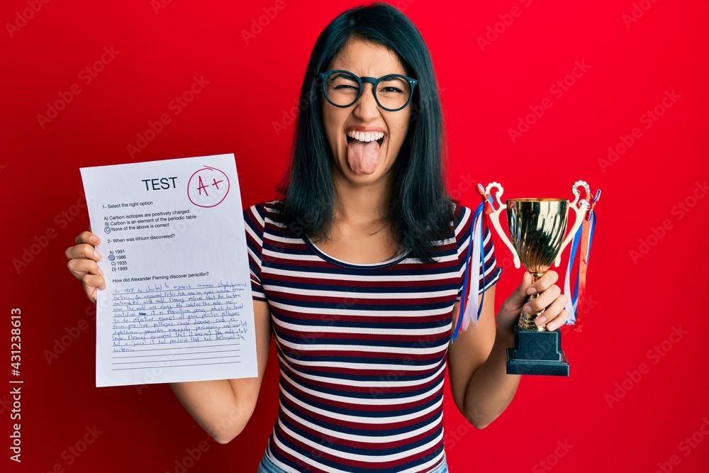 Beautiful asian young woman showing a passed exam holding trophy sticking tongue out happy with funny expression. Beautiful asian young woman showing a passed exam holding trophy sticking tongue out happy with funny expression.