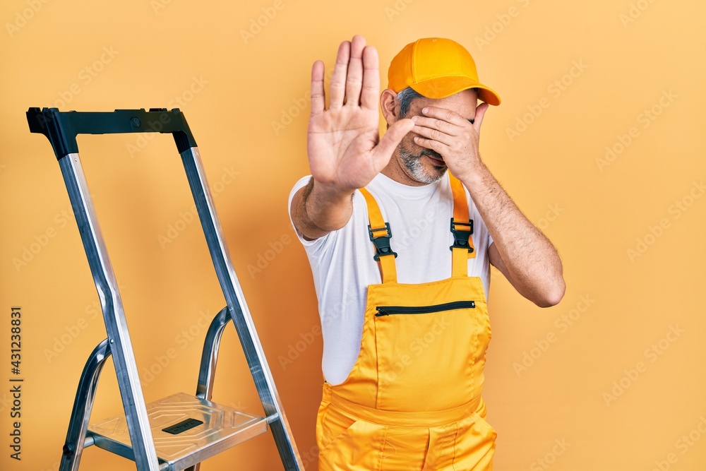Handsome middle age man with grey hair holding ladder covering eyes with hands and doing stop gesture with sad and fear expression. embarrassed and negative concept. Handsome middle age man with grey hair holding ladder covering eyes with hands and doing stop gesture with sad and fear expression. embarrassed and negative concept.
