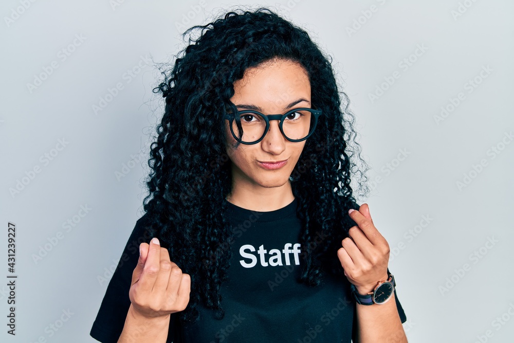 Young hispanic woman with curly hair wearing staff t shirt doing money gesture with hands, asking for salary payment, millionaire business Young hispanic woman with curly hair wearing staff t shirt doing money gesture with hands, asking for salary payment, millionaire business