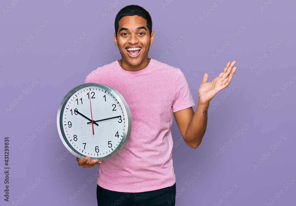 Young handsome hispanic man holding big clock celebrating victory with happy smile and winner expression with raised hands Young handsome hispanic man holding big clock celebrating victory with happy smile and winner expression with raised hands