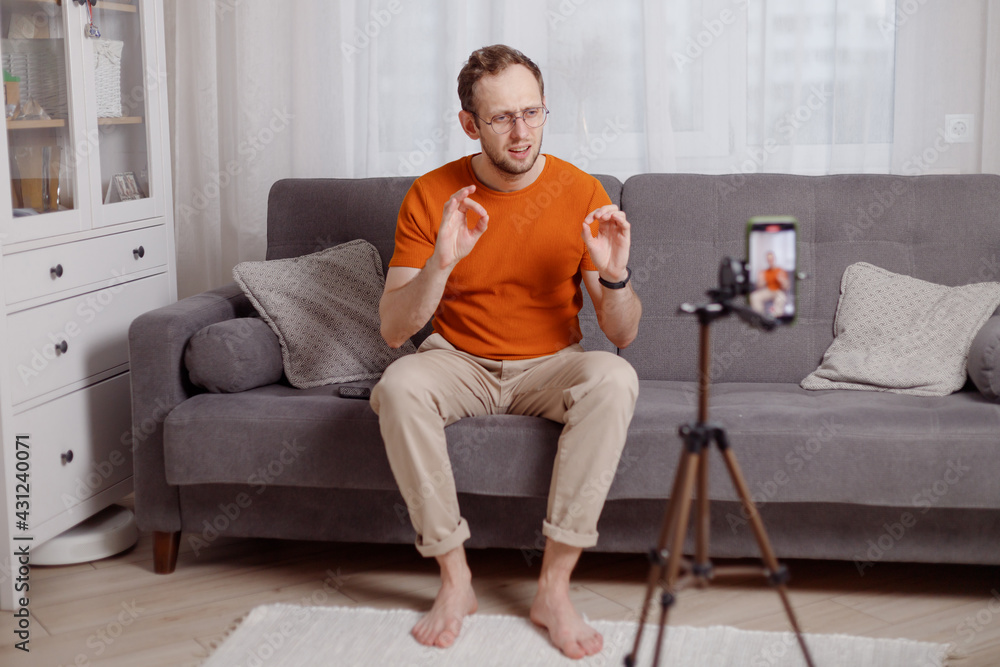 Fototapeta premium Hipster man blogger recording vlog on mobile phone sit on couch in living room and explain and gesturing with hands