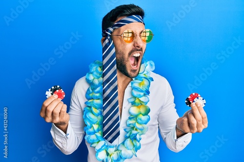 Young hispanic businessman wearing party funny style holding casino chips angry and mad screaming frustrated and furious, shouting with anger. rage and aggressive concept. photo