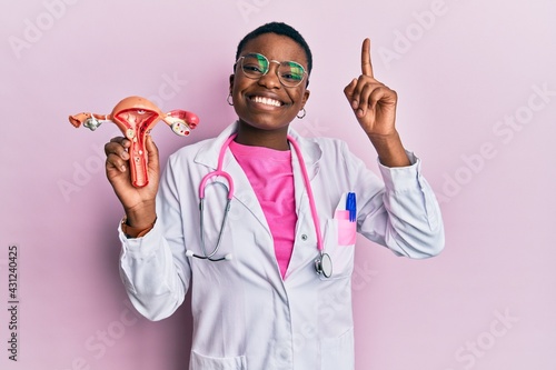 Young african american doctor woman holding anatomical model of female genital organ smiling with an idea or question pointing finger with happy face, number one photo