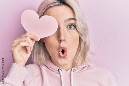 Young blonde girl holding heart shape paper over eye scared and amazed with open mouth for surprise, disbelief face