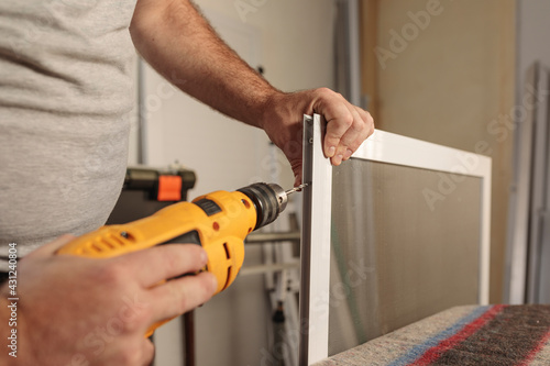 Man's hand with a drill to assemble an aluminum window screen. Selective focus. Horizontal image.