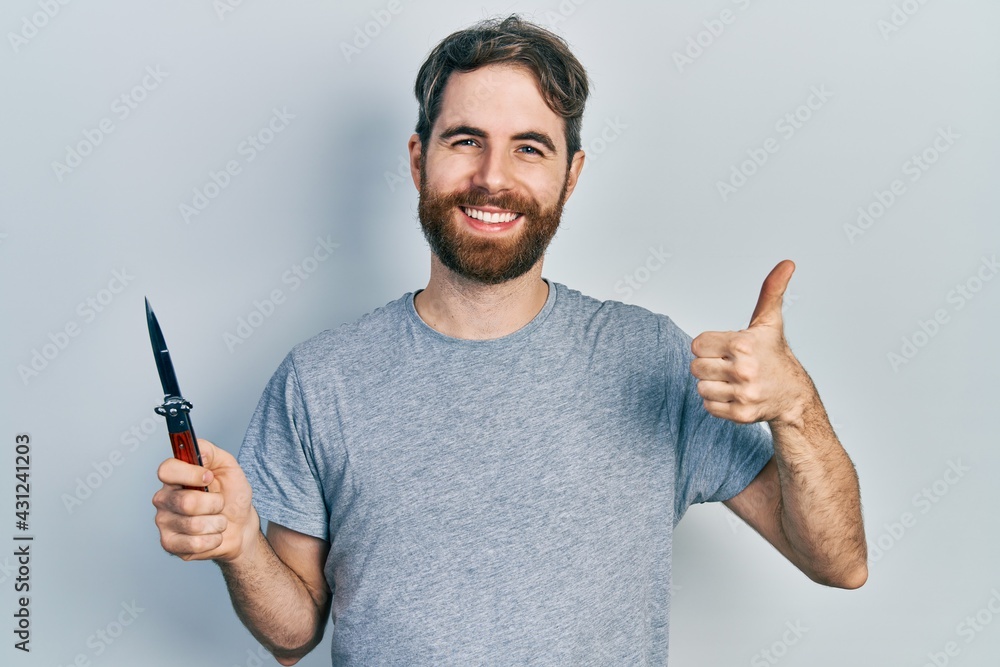 Caucasian man with beard holding pocket knife smiling happy and positive, thumb up doing excellent and approval sign Caucasian man with beard holding pocket knife smiling happy and positive, thumb up doing excellent and approval sign