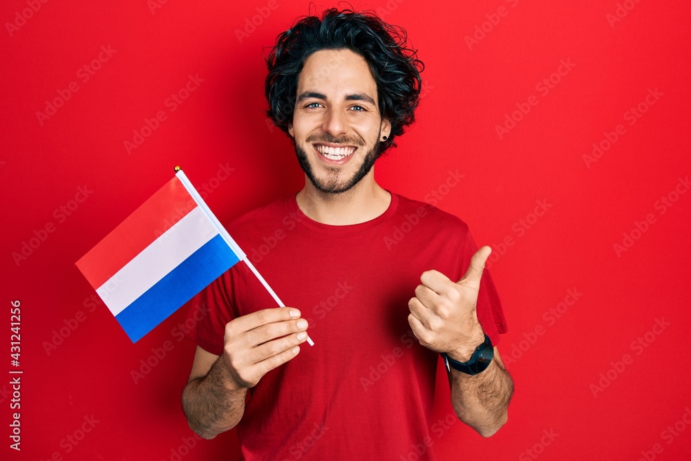 Handsome hispanic man holding netherlands flag smiling happy and positive, thumb up doing excellent and approval sign Handsome hispanic man holding netherlands flag smiling happy and positive, thumb up doing excellent and approval sign