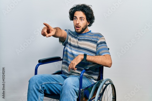 Handsome hispanic man sitting on wheelchair pointing with finger surprised ahead, open mouth amazed expression, something on the front