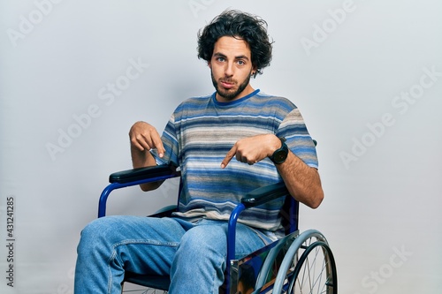 Handsome hispanic man sitting on wheelchair pointing down looking sad and upset, indicating direction with fingers, unhappy and depressed.