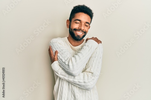 Handsome hispanic man with beard wearing casual winter sweater hugging oneself happy and positive, smiling confident. self love and self care