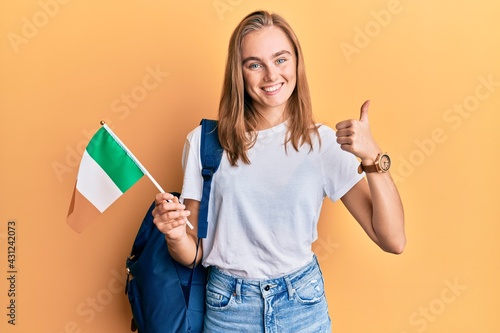 Beautiful blonde woman exchange student holding ireland flag smiling happy and positive, thumb up doing excellent and approval sign