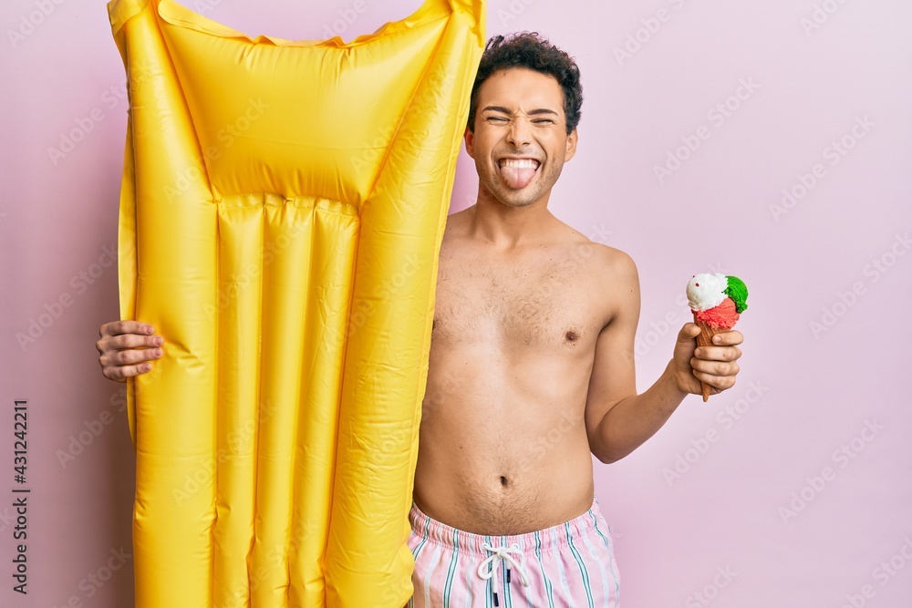 Obraz premium Young handsome man wearing swimsuit holding summer mattress float and icecream sticking tongue out happy with funny expression.
