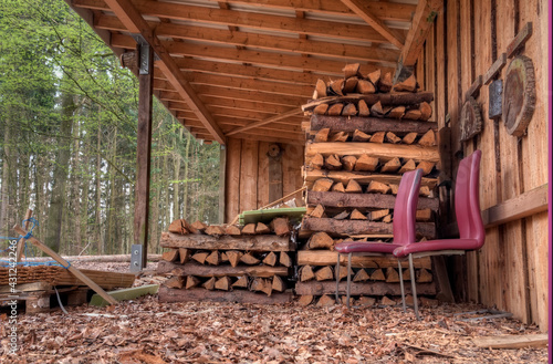 Close-up of a warehouse for lumber and lumberjack tools in a wood. Sheltered under the roof are two red leather chairs and piles of chopped wood.
Perspective view from the ground.
