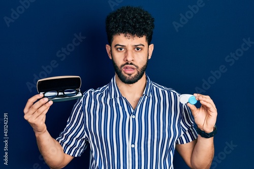 Young arab man with beard holding glasses and contact lenses in shock face, looking skeptical and sarcastic, surprised with open mouth