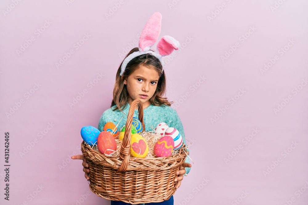 Little beautiful girl wearing cute easter bunny ears holding wicker basket with colored eggs clueless and confused expression. doubt concept. Little beautiful girl wearing cute easter bunny ears holding wicker basket with colored eggs clueless and confused expression. doubt concept.
