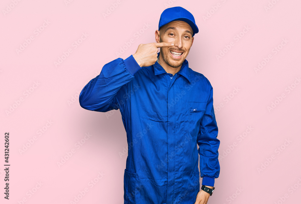 Bald man with beard wearing builder jumpsuit uniform pointing with hand finger to face and nose, smiling cheerful. beauty concept