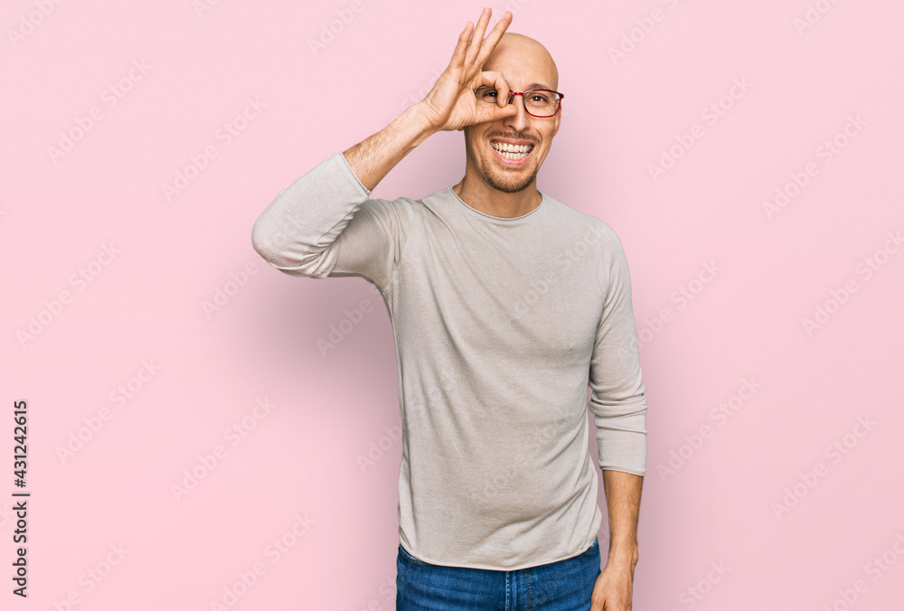 Bald man with beard wearing casual clothes and glasses doing ok gesture with hand smiling, eye looking through fingers with happy face.