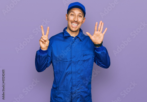 Bald man with beard wearing builder jumpsuit uniform showing and pointing up with fingers number seven while smiling confident and happy.