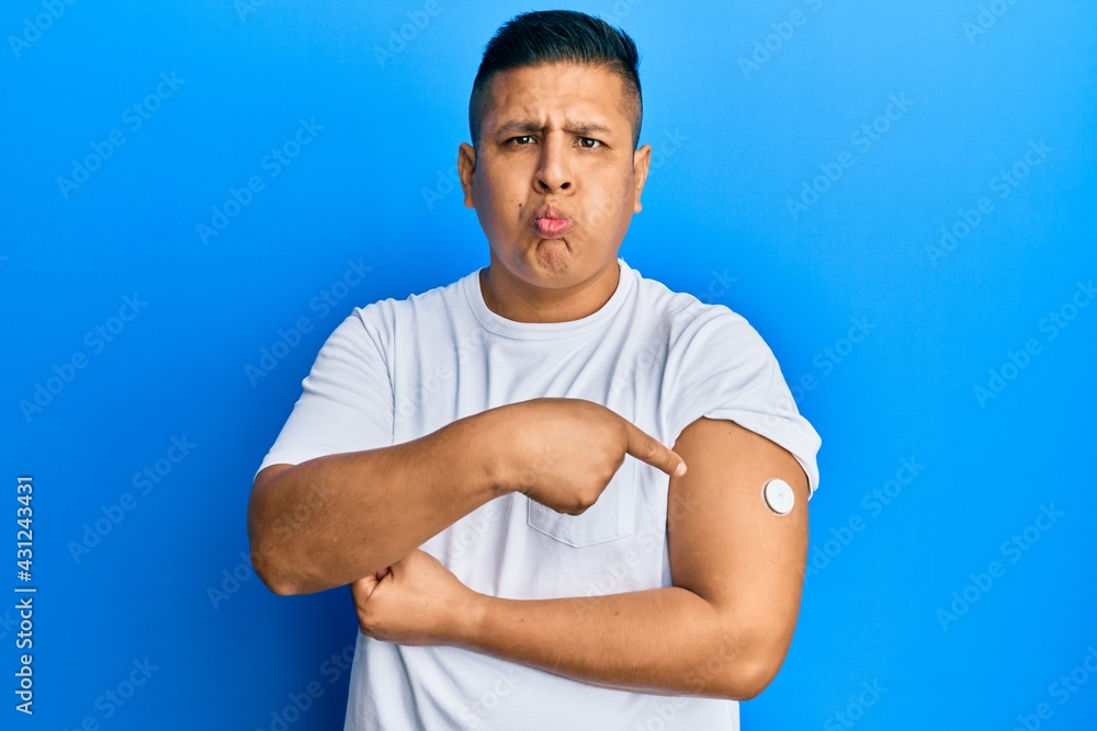 Young latin man using glucose meter button on arm puffing cheeks with funny face. mouth inflated with air, catching air. Young latin man using glucose meter button on arm puffing cheeks with funny face. mouth inflated with air, catching air.