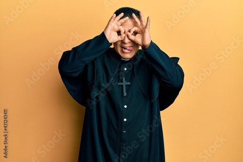 Young latin priest man standing over yellow background doing ok gesture like binoculars sticking tongue out, eyes looking through fingers. crazy expression. © Krakenimages.com