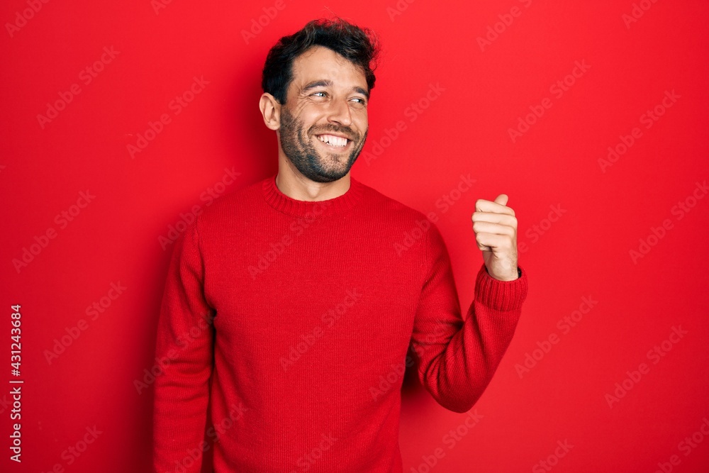 Fototapeta premium Handsome man with beard wearing casual red sweater smiling with happy face looking and pointing to the side with thumb up.