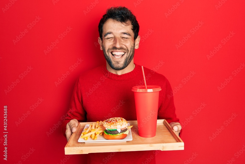 Handsome man with beard eating a tasty classic burger with fries and soda smiling and laughing hard out loud because funny crazy joke. Handsome man with beard eating a tasty classic burger with fries and soda smiling and laughing hard out loud because funny crazy joke.