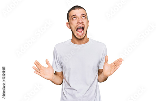Hispanic young man wearing casual white t shirt crazy and mad shouting and yelling with aggressive expression and arms raised. frustration concept.