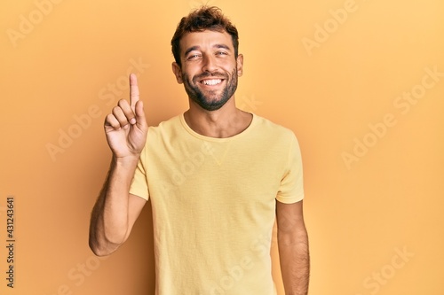 Handsome man with beard wearing casual yellow tshirt over yellow background showing and pointing up with finger number one while smiling confident and happy.