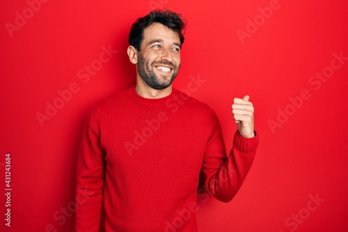 Handsome man with beard wearing casual red sweater smiling with happy face looking and pointing to the side with thumb up. © Krakenimages.com