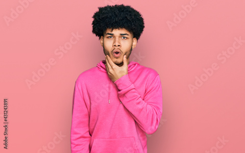 Young african american man with afro hair wearing casual pink sweatshirt looking fascinated with disbelief, surprise and amazed expression with hands on chin
