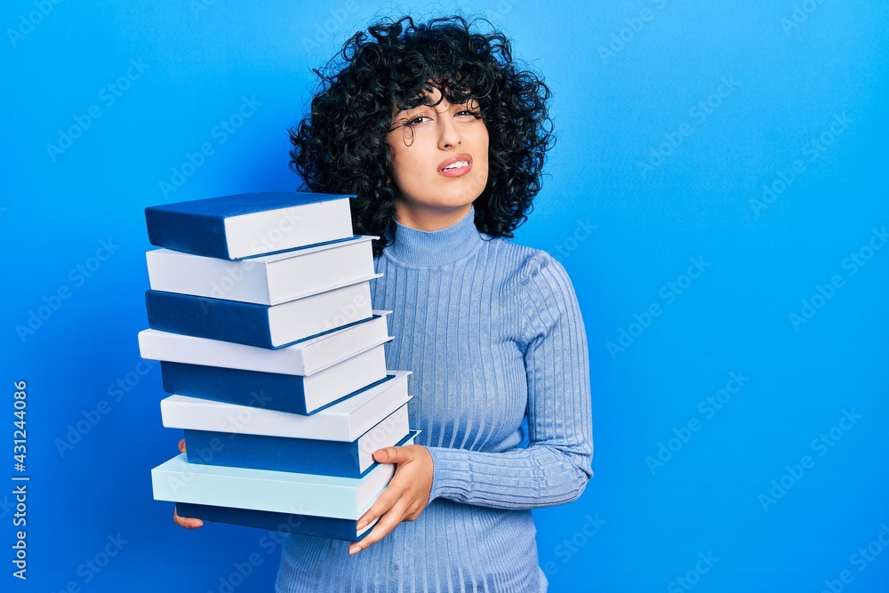 Young middle east woman holding a pile of books clueless and confused expression. doubt concept. Young middle east woman holding a pile of books clueless and confused expression. doubt concept.