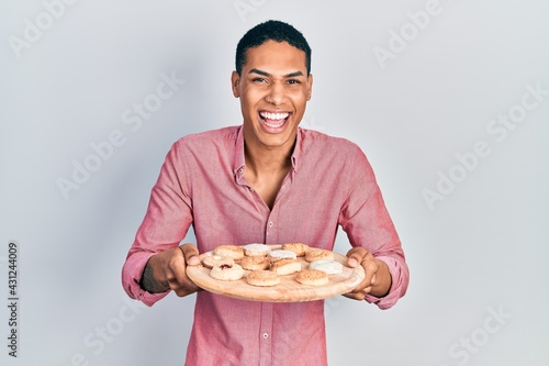 Young african american guy holding tray with cake sweets smiling and laughing hard out loud because funny crazy joke.