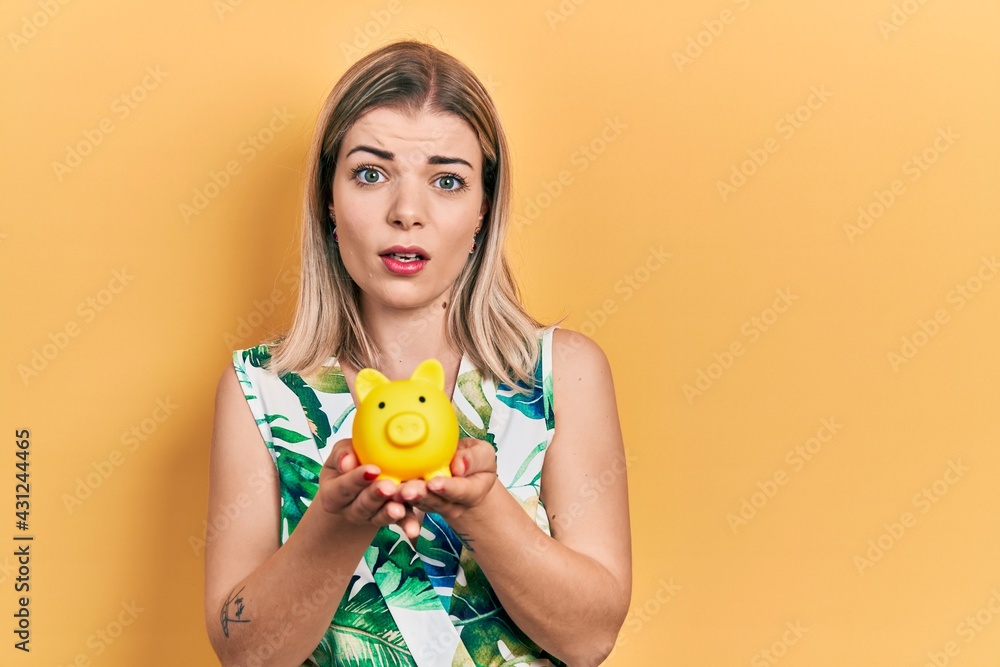 Beautiful caucasian woman holding piggy bank clueless and confused expression. doubt concept. Beautiful caucasian woman holding piggy bank clueless and confused expression. doubt concept.