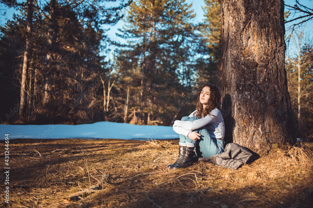 Young, brunette girl sitting in the forest, leaning on the tree