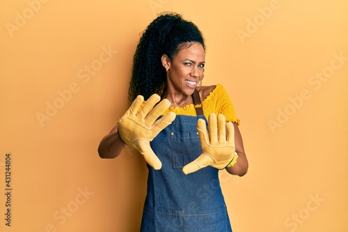 Middle age african american woman wearing professional apron afraid and terrified with fear expression stop gesture with hands, shouting in shock. panic concept.
