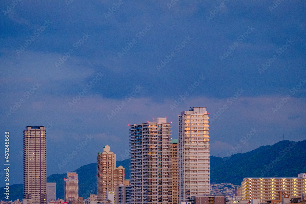 兵庫県神戸市の都市景観。六甲山系を背景に高層タワーマンションが立ち並ぶ。朝日に照らされてビルが輝く。