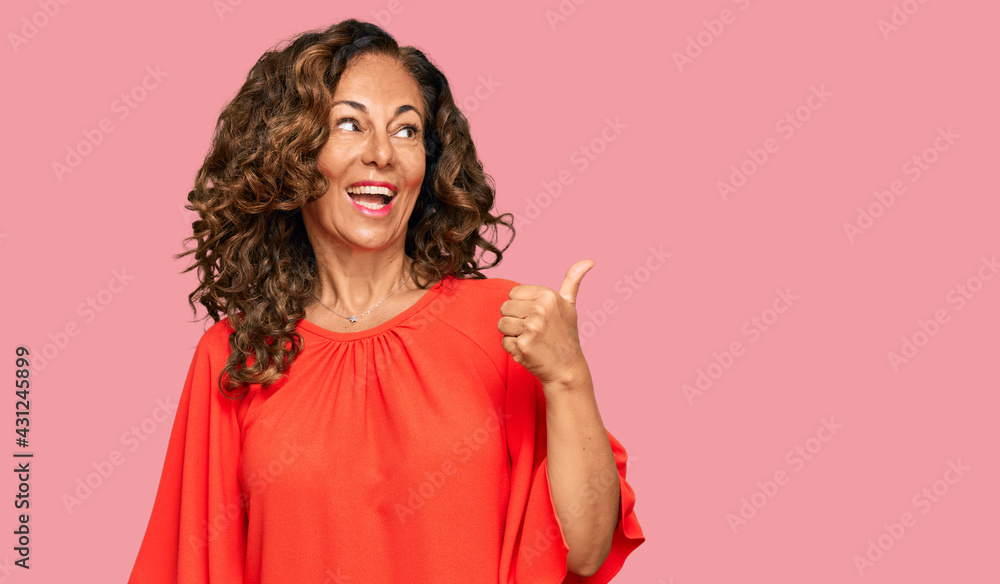 Middle age hispanic woman wearing casual clothes smiling with happy face looking and pointing to the side with thumb up. Middle age hispanic woman wearing casual clothes smiling with happy face looking and pointing to the side with thumb up.