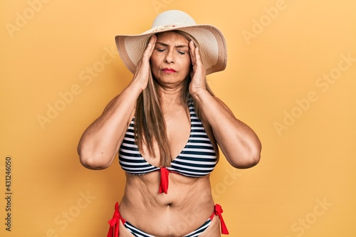 Middle age hispanic woman wearing bikini and summer hat with hand on head for pain in head because stress. suffering migraine.