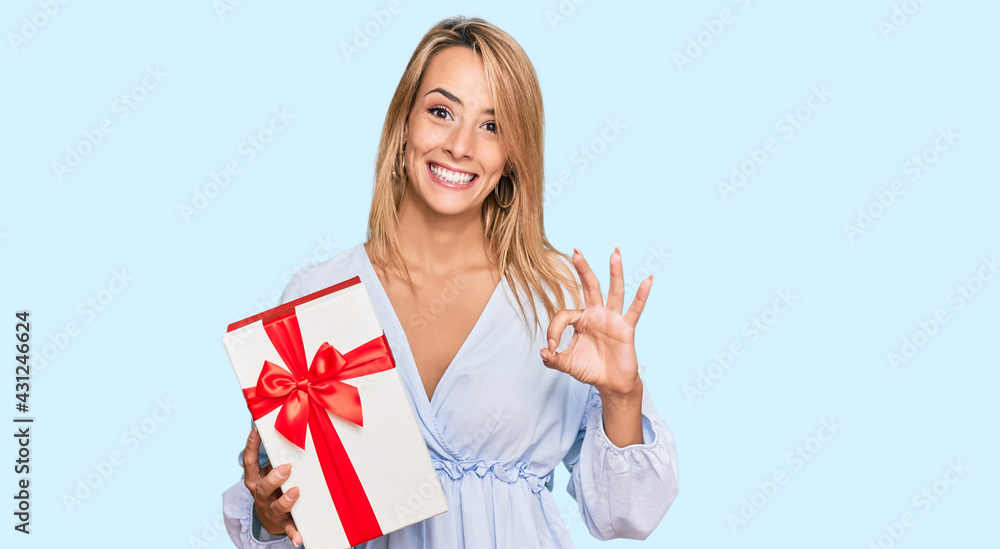 Beautiful blonde woman holding gift doing ok sign with fingers, smiling friendly gesturing excellent symbol Beautiful blonde woman holding gift doing ok sign with fingers, smiling friendly gesturing excellent symbol