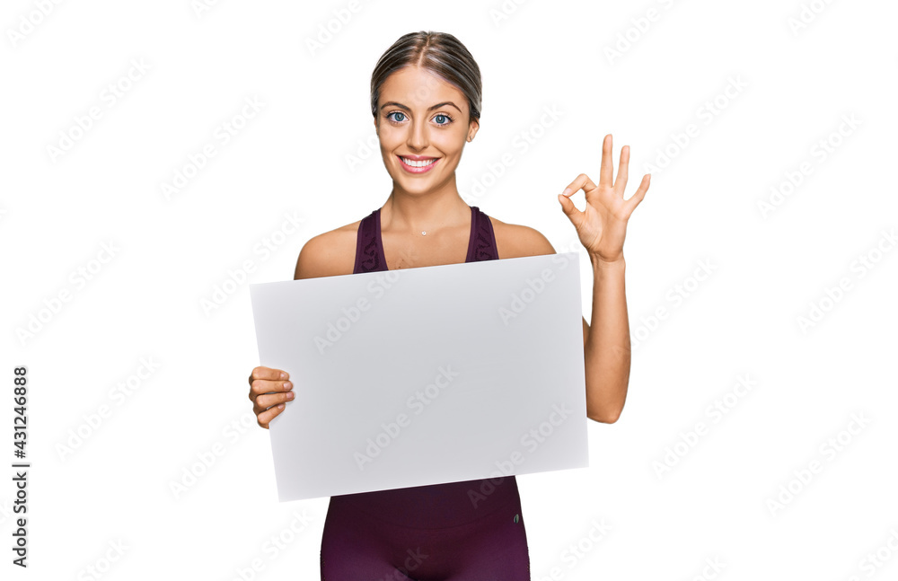 Beautiful blonde woman wearing sportswear holding white banner doing ok sign with fingers, smiling friendly gesturing excellent symbol Beautiful blonde woman wearing sportswear holding white banner doing ok sign with fingers, smiling friendly gesturing excellent symbol