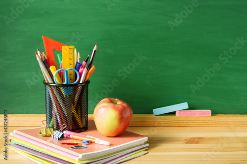 School supplies, pens, pencils in a glass, notebooks, chalk for a blackboard and an apple on a wooden table against the background of a green school board, copy space.  School background