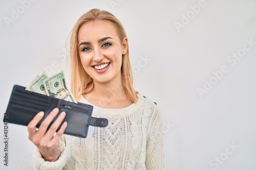Young blonde girl smiling happy holding wallet with dollars standing at the city.
