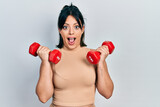 Young hispanic woman wearing sportswear using dumbbells afraid and shocked with surprise and amazed expression, fear and excited face.
