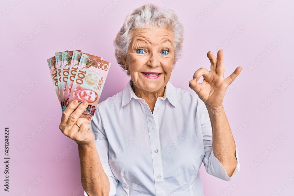 Senior grey-haired woman holding 100 new zealand dollars banknote doing ok sign with fingers, smiling friendly gesturing excellent symbol Senior grey-haired woman holding 100 new zealand dollars banknote doing ok sign with fingers, smiling friendly gesturing excellent symbol