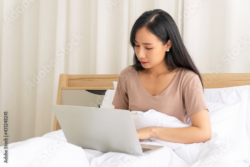 Freelance young woman using laptop working on internet write something on the book while sitting on white bed with pillow in the morning at apartment. Lifestyle work at home with technology concept.