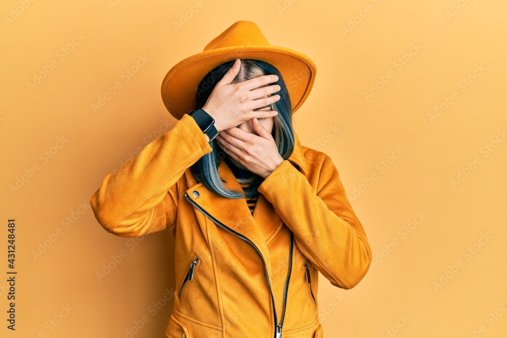 Young modern girl wearing yellow hat and leather jacket covering eyes and mouth with hands, surprised and shocked. hiding emotion Young modern girl wearing yellow hat and leather jacket covering eyes and mouth with hands, surprised and shocked. hiding emotion